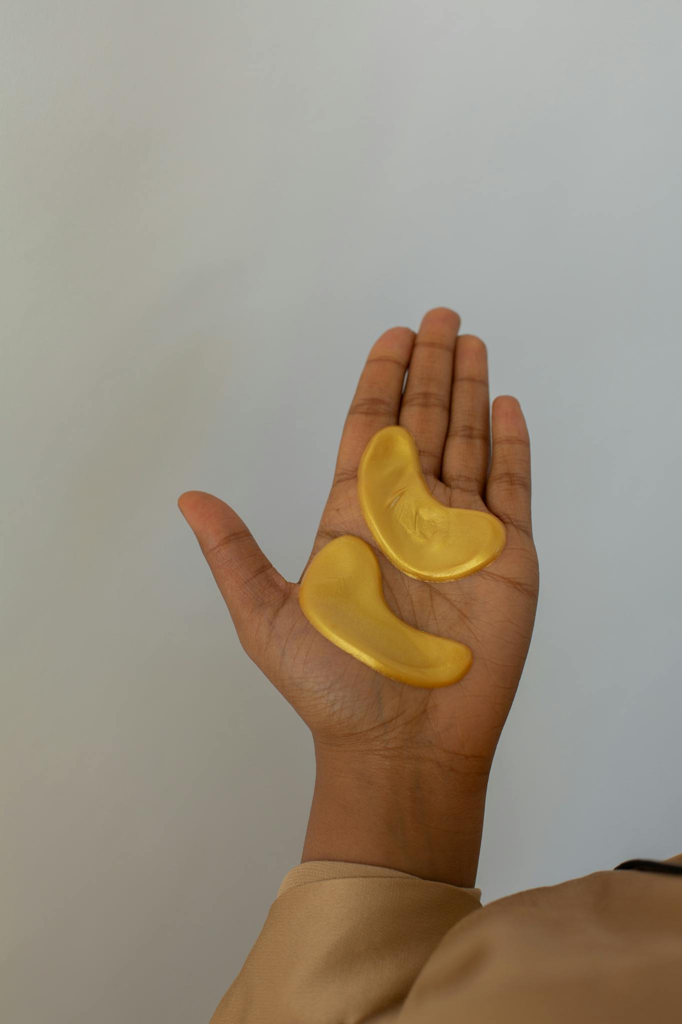 A hand holding gold eye masks against a neutral background, emphasizing skincare and relaxation.