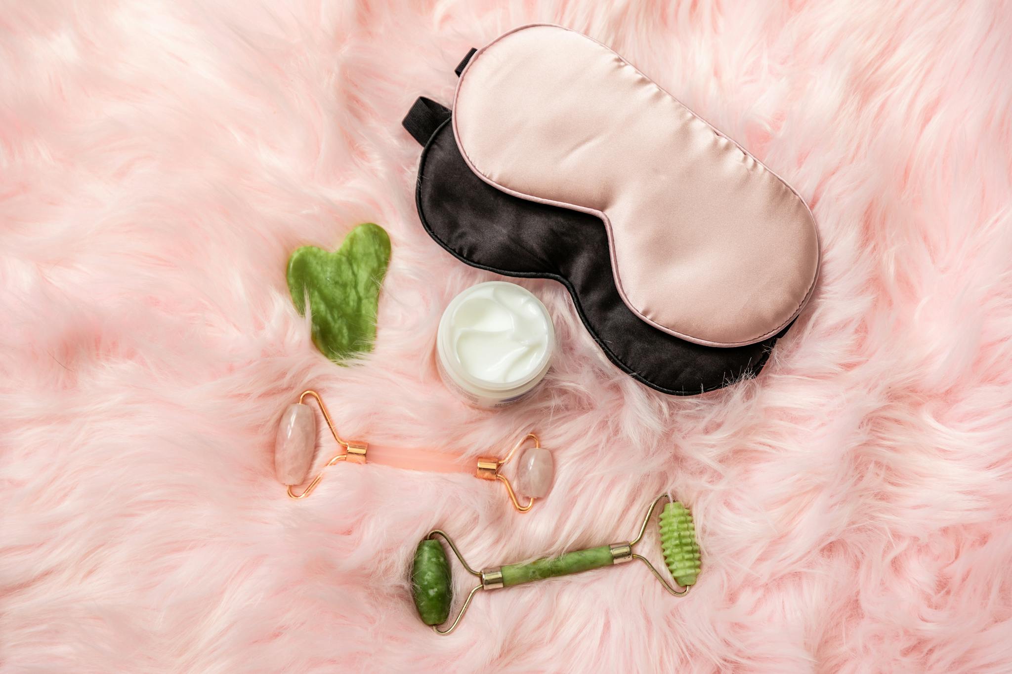 Flat lay of eye mask, jade roller, cream, and more on soft pink fur.
